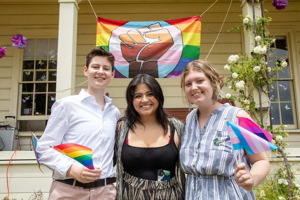 Three people standing in front of the LBGTQ+  flag